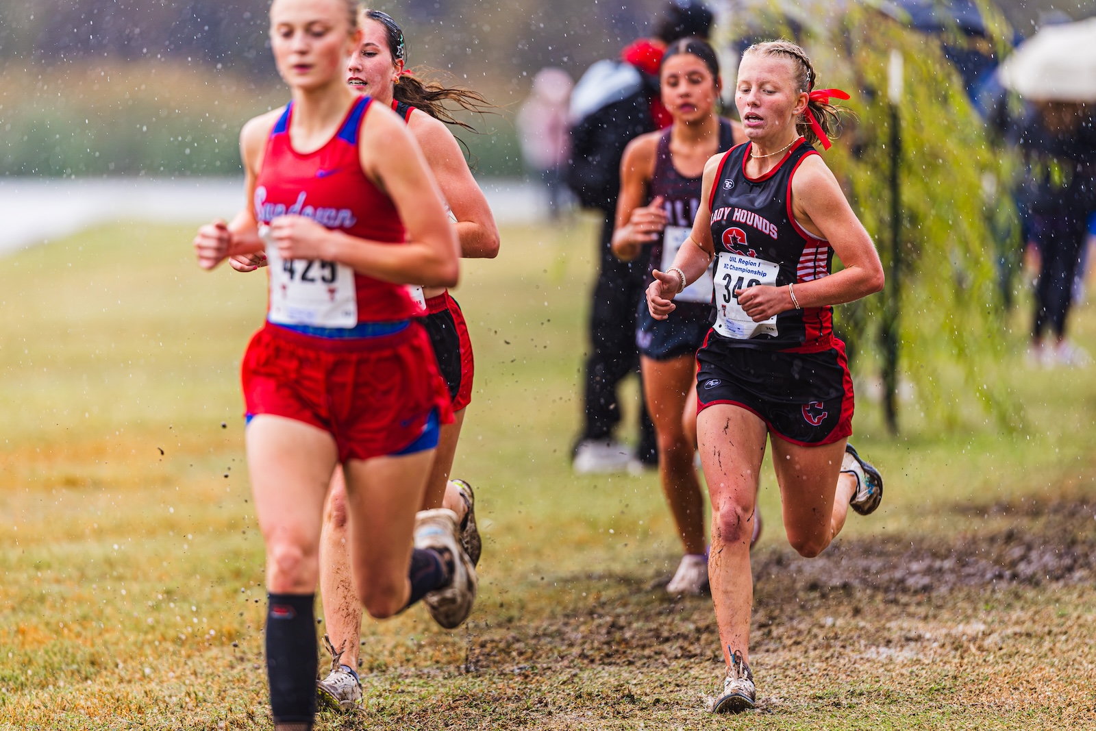 DE3_7440 | Press Pass Sports Female runners in red and black uniforms sprint through a muddy, rainy cross-country course. (Informative)