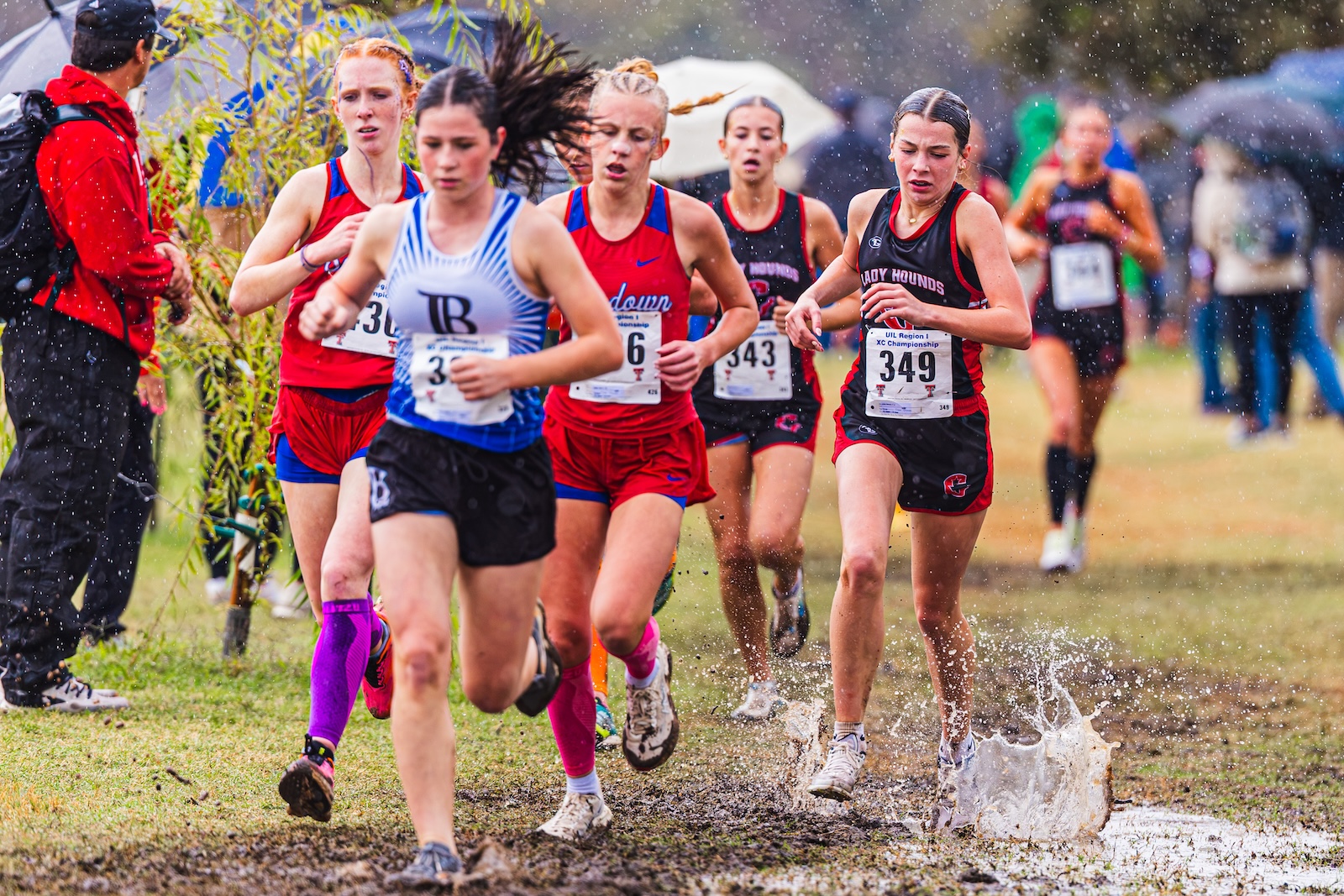 DE3_7462 | Press Pass Sports Female runners in a cross-country race sprint through a muddy puddle in the rain, wearing numbered bibs and colorful uniforms.