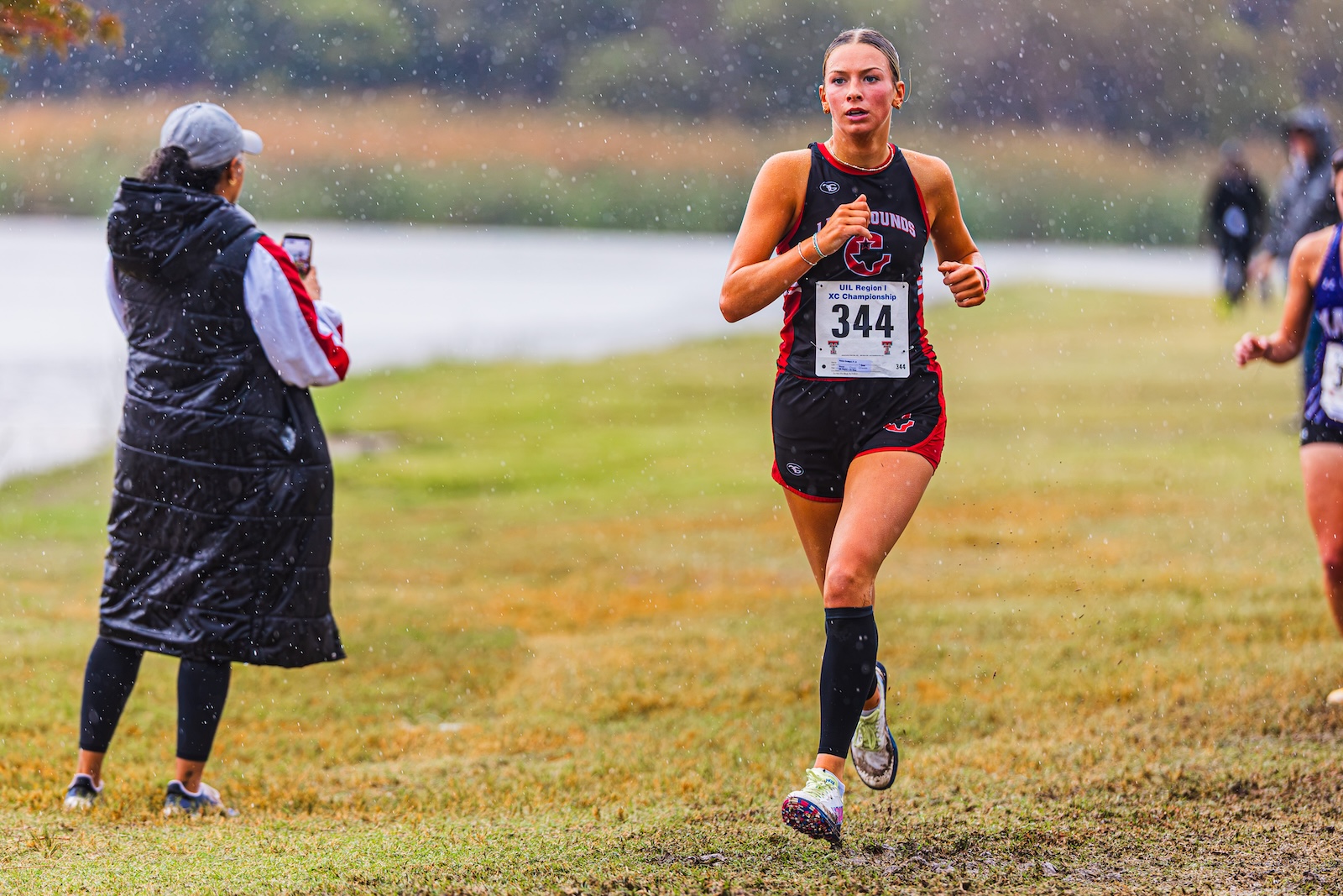 DE3_7502 | Press Pass Sports Female runner wearing bib 344 races through a muddy grassy field in light rain, while a spectator films from the left.