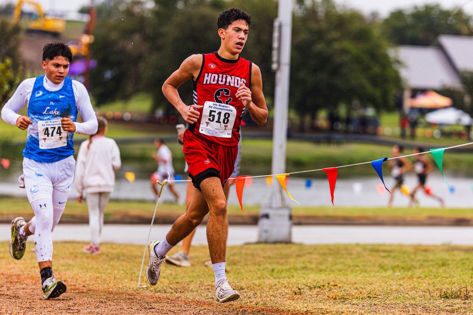 DE3_7990 | Press Pass Sports Two young runners race on a wet track; foreground is a sprinter in a red HOUNDS uniform (bib 518) leading, blue-uniform rival (#474) nearby. colorful pennant markers line the course.
