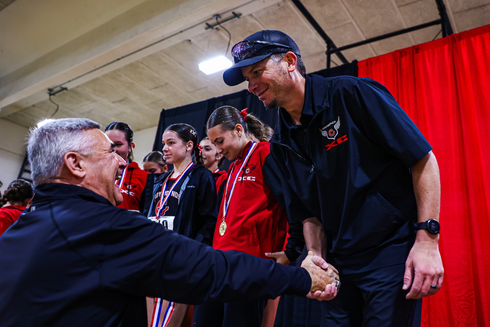 DEP29649 | Press Pass Sports Coach shakes hands with a medal-winning athlete on stage during an awards ceremony, red curtain in the background.