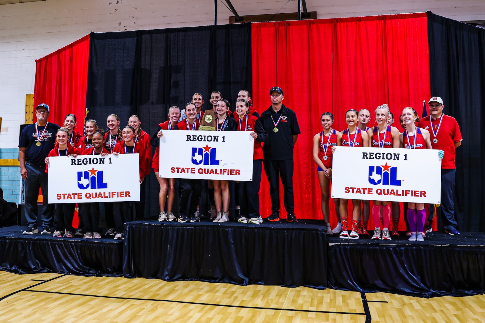 DEP29833 | Press Pass Sports Group of young athletes on a stage holding 'REGION 1 UIL STATE QUALIFIER' banners and a trophy, wearing red uniforms and medals, with coaches nearby.