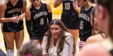 Girls' basketball team in black Amarillo jerseys form a huddle around their coach on a gym floor during a timeout.