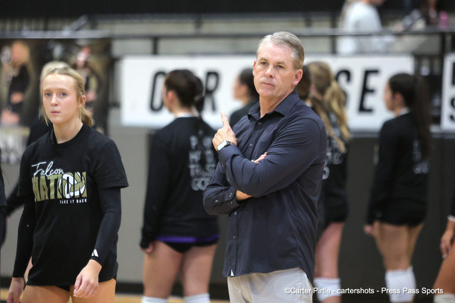 A coach in a dark shirt stands with arms crossed, pointing a finger while athletes practice in the background in a gym.