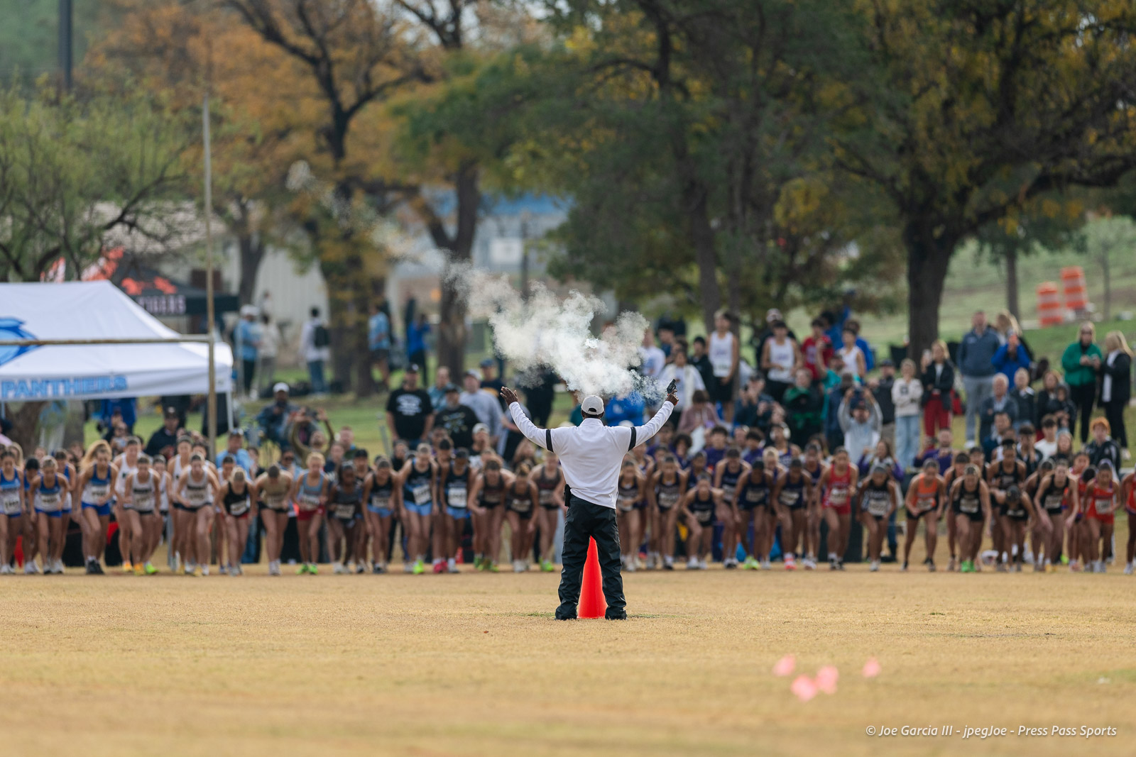 regional-xc-lubbock-2026-jpegJoe-pps-1 | Press Pass Sports Athletes lined up at the starting line of a race as the official raises arms and white smoke erupts from a starting pistol, spectators in the background.