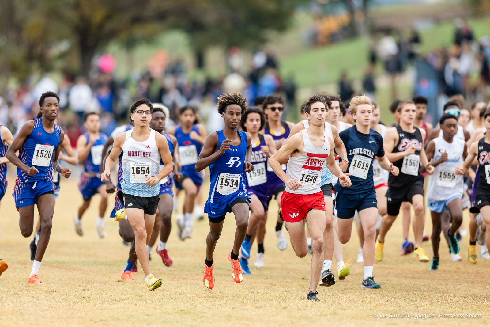 regional-xc-lubbock-2026-jpegJoe-pps-4 | Press Pass Sports Group of runners sprinting in a cross-country race on a grassy field, bib numbers visible and crowds in the background.