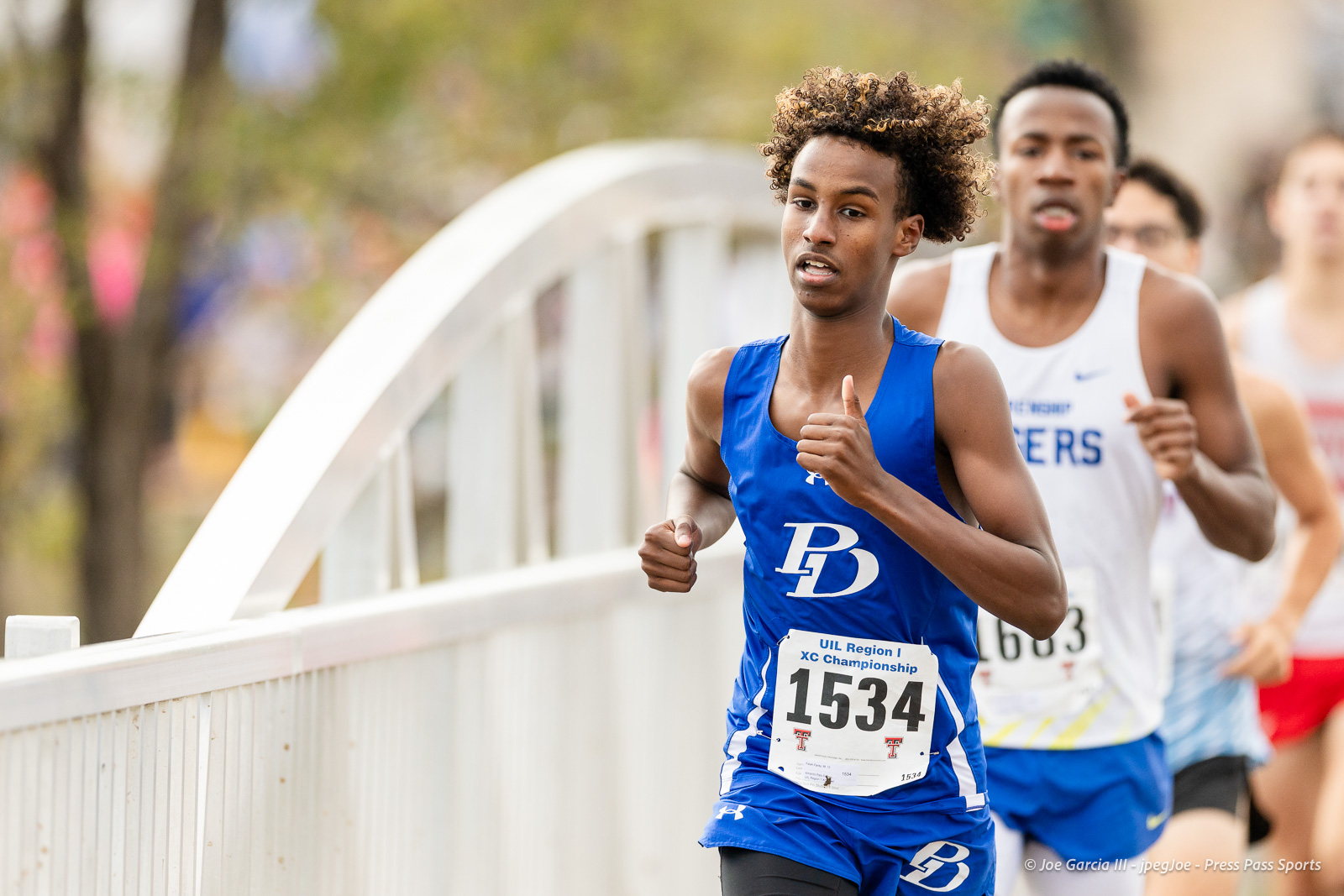regional-xc-lubbock-2026-jpegJoe-pps-5 | Press Pass Sports Male runner in blue uniform with bib 1534 leading a race on a curved bridge, others follow closely behind on a sunny day.