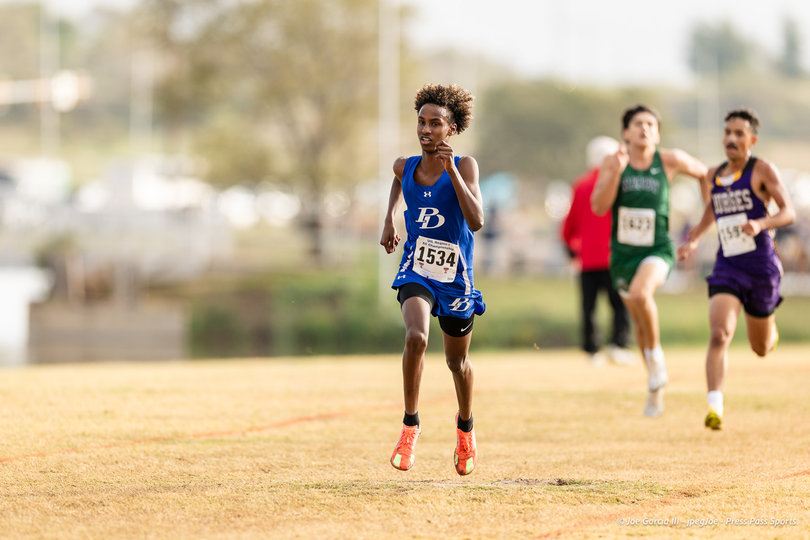 regional-xc-lubbock-2026-jpegJoe-pps-6 | Press Pass Sports Young male sprinter in a blue uniform (bib 1534) leads a race on a grassy track with others behind him in green and purple jerseys, under sunny skies.