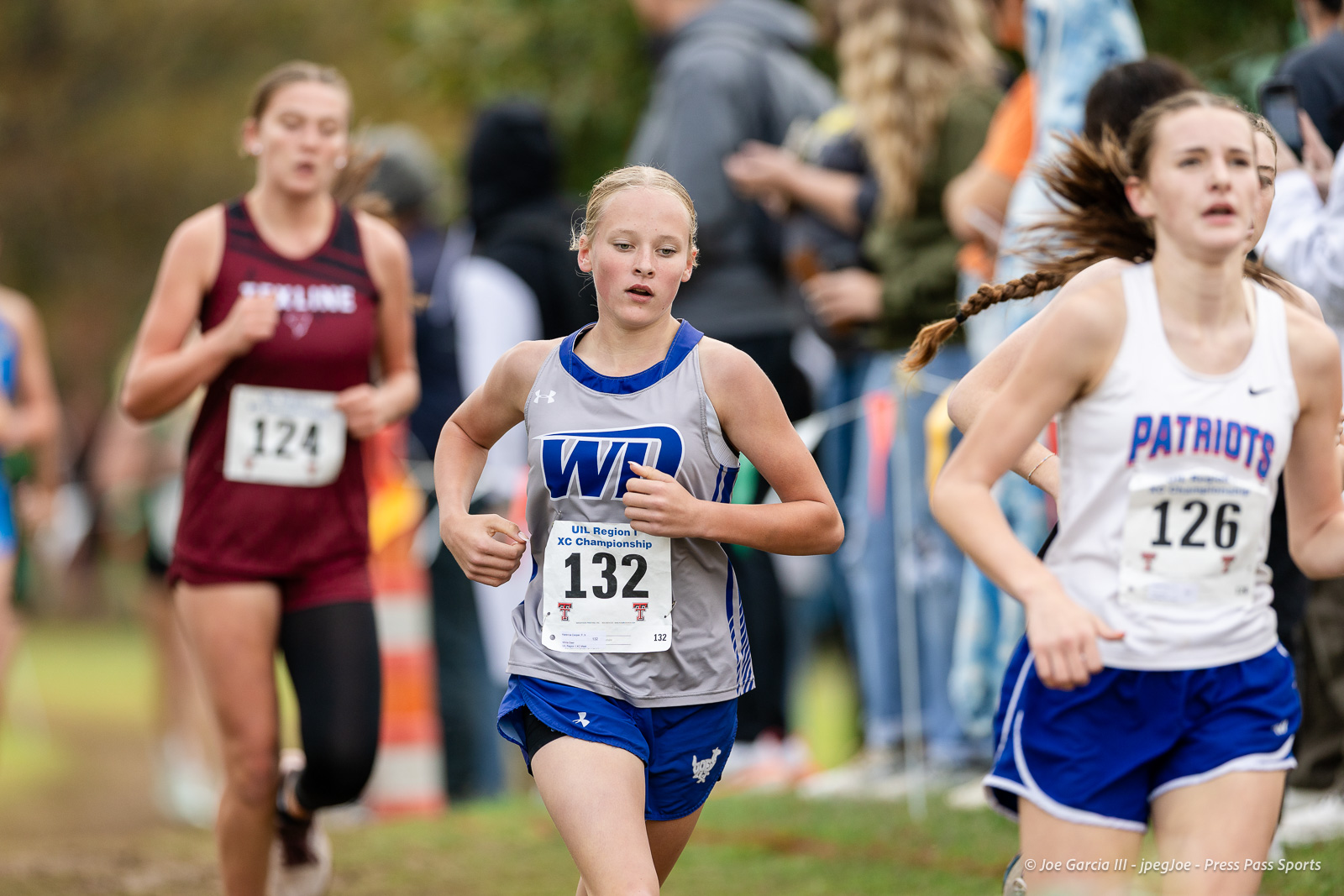 regional-xc-lubbock-2026-jpegJoe-pps-9 | Press Pass Sports Young female runner in gray-blue uniform (132) leading a cross-country race, others behind on a grassy course with spectators nearby