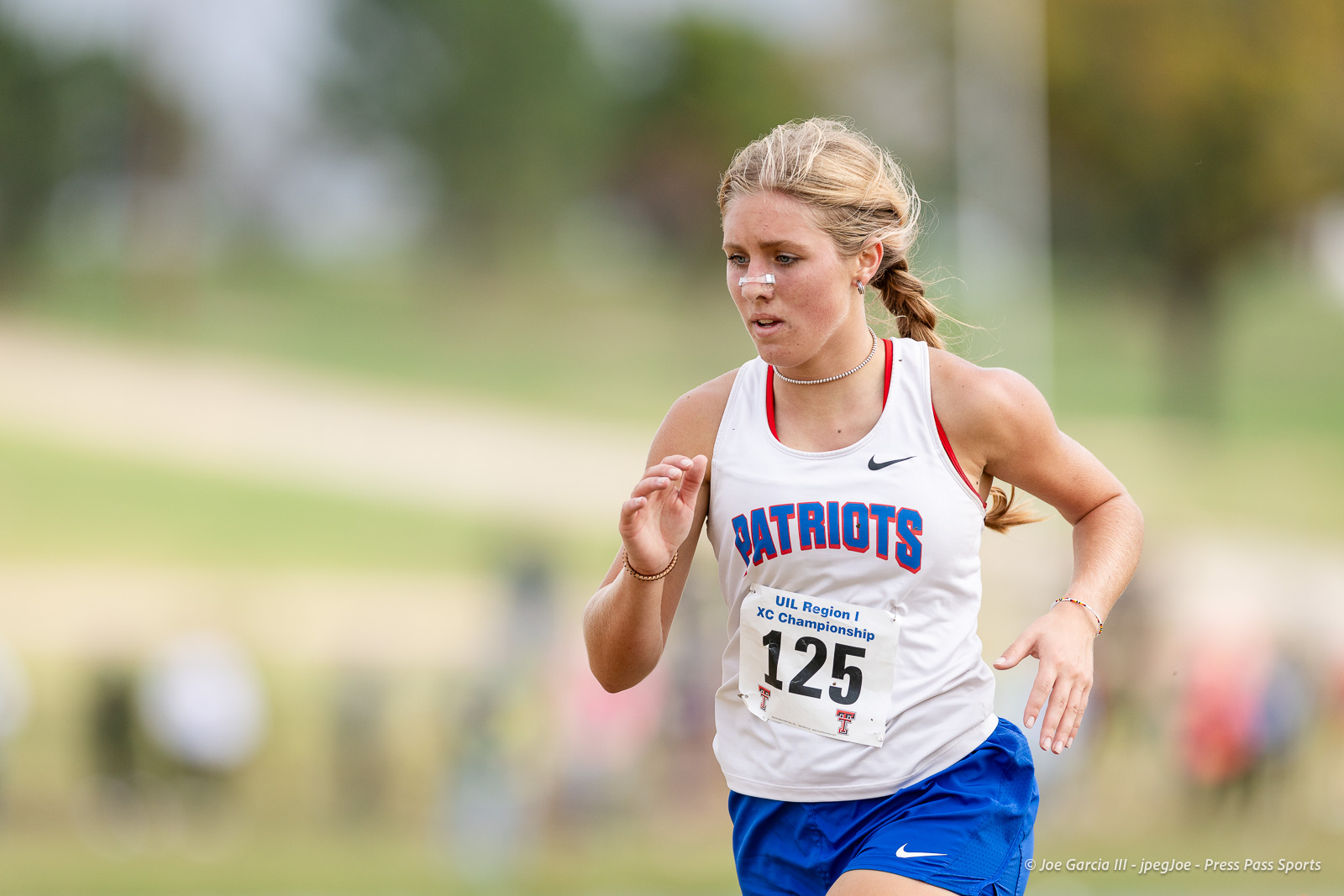 regional-xc-lubbock-2026-jpegJoe-pps-11 | Press Pass Sports Female athlete running cross-country in a Patriots jersey with race bib 125, outdoors on a blurred course field.