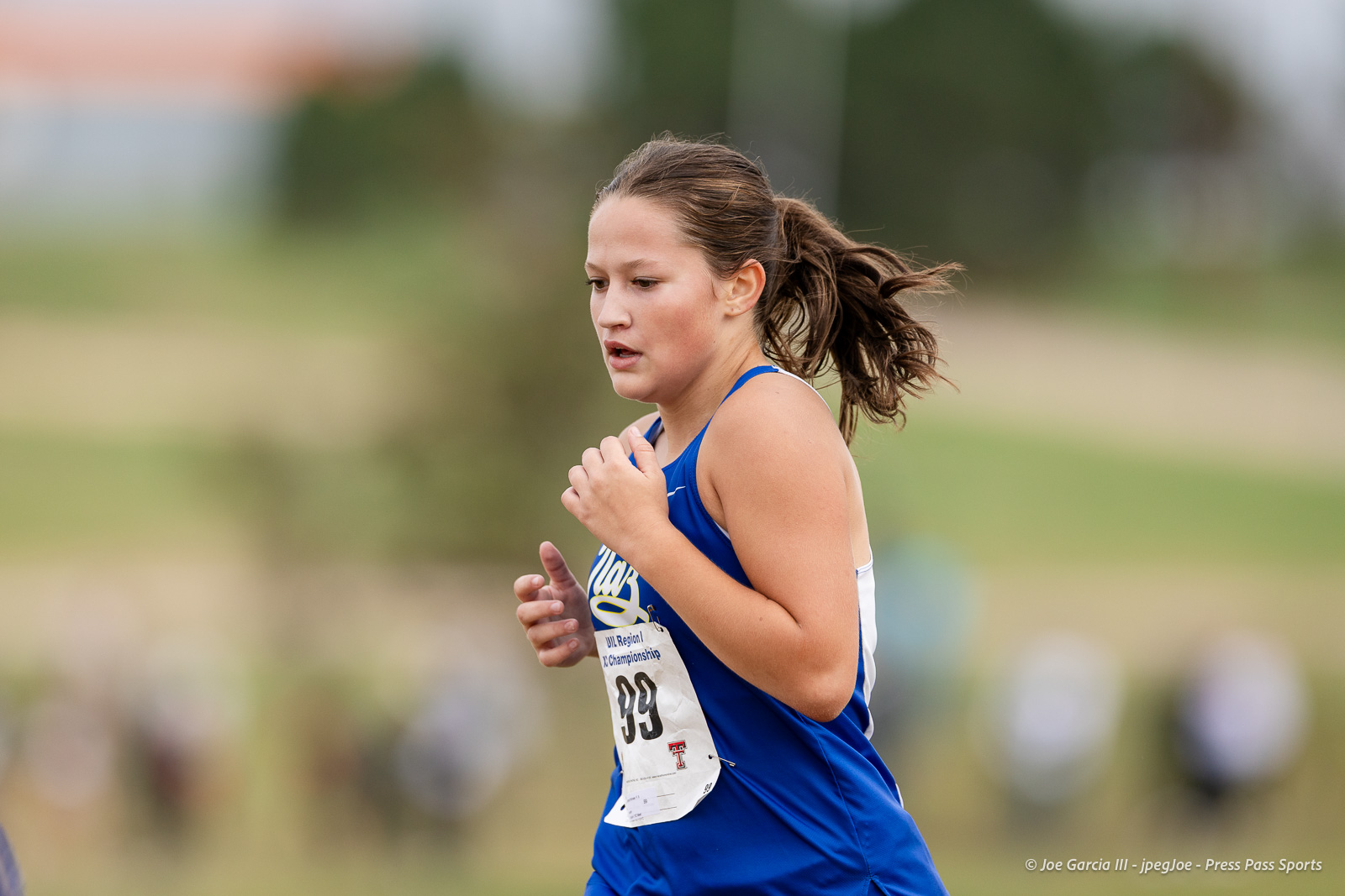 regional-xc-lubbock-2026-jpegJoe-pps-12 | Press Pass Sports Female runner in a blue singlet with bib 99 sprinting in a race on a grassy field, focused expression.