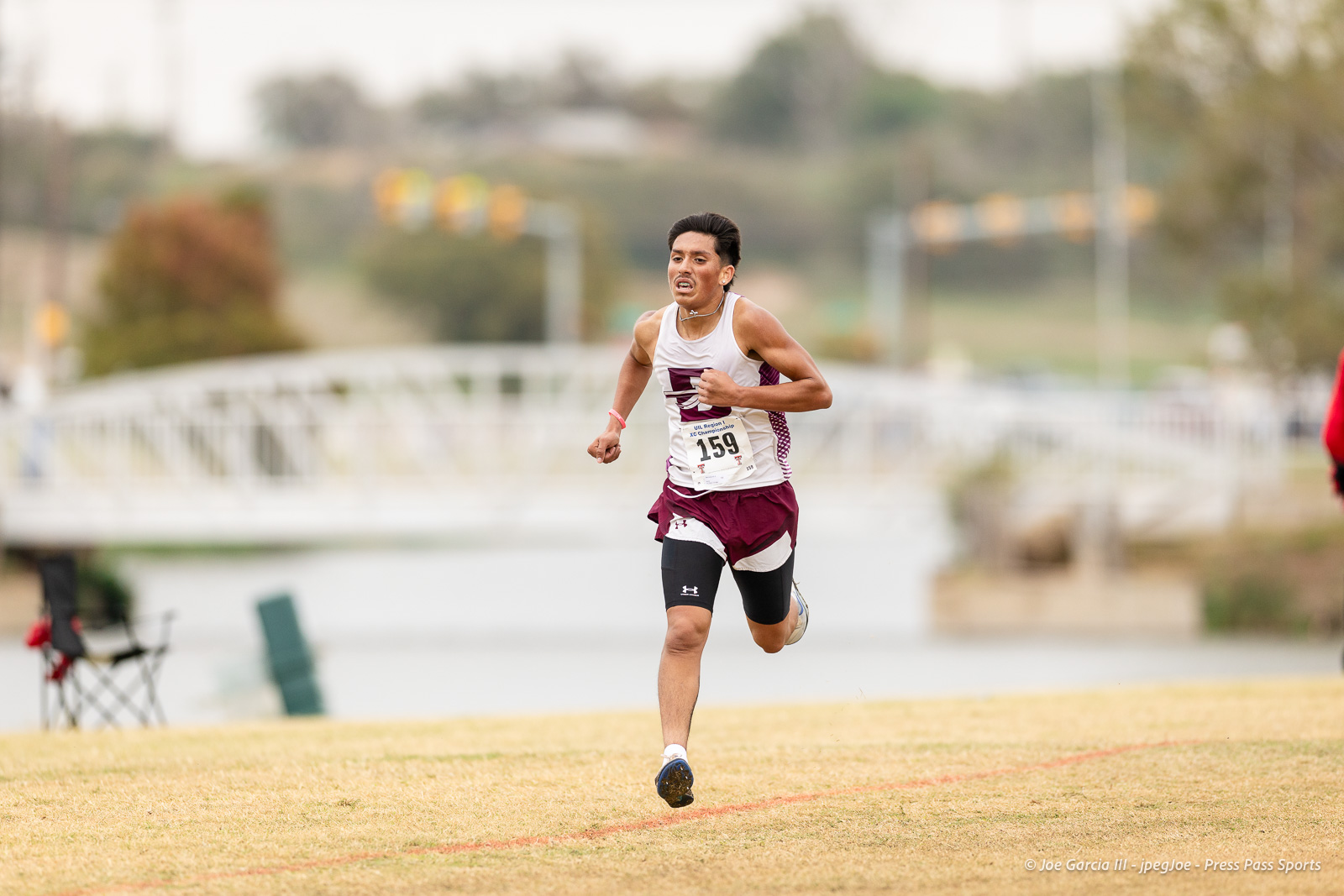 regional-xc-lubbock-2026-jpegJoe-pps-14 | Press Pass Sports Male runner wearing a white and maroon tank top and shorts, bib 159, sprinting on a grassy field during a race with a blurred background.