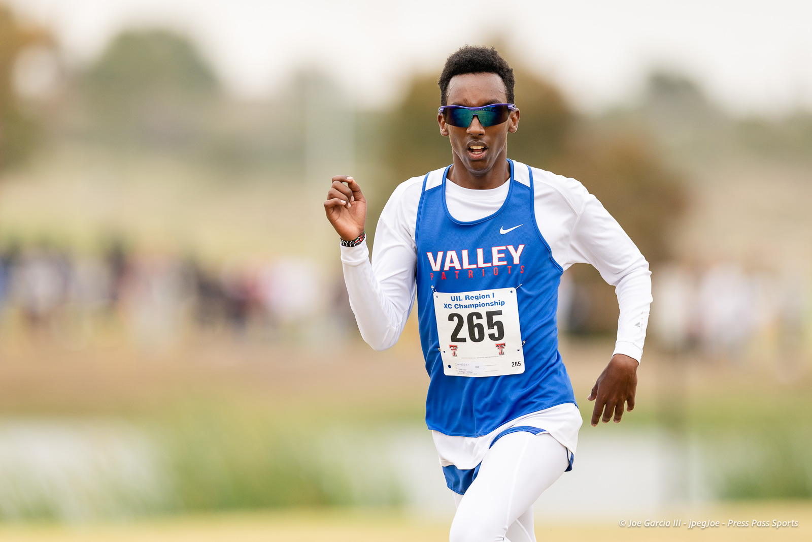 regional-xc-lubbock-2026-jpegJoe-pps-15 | Press Pass Sports Male cross-country runner in a blue Valley Patriots singlet, white long sleeves, black sunglasses, mid-stride during a race; bib number 265 visible.