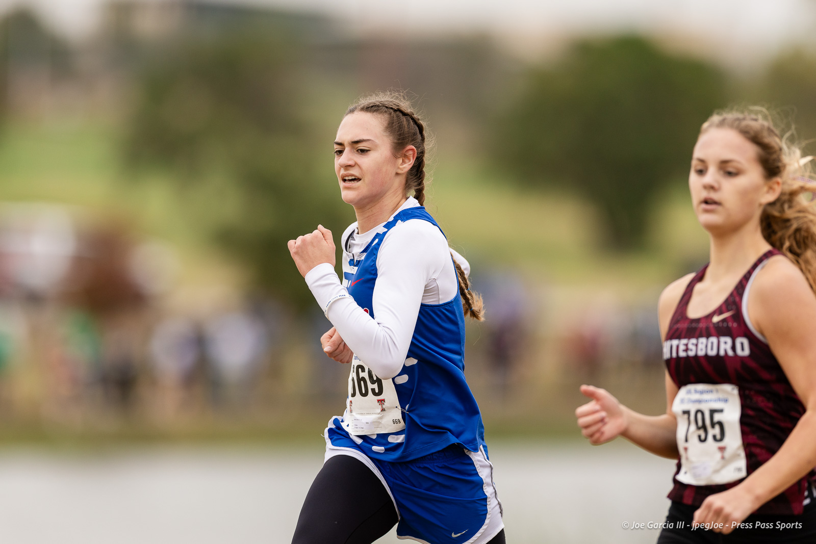 regional-xc-lubbock-2026-jpegJoe-pps-18 | Press Pass Sports Two female runners compete in an outdoor race; the runner in blue leads while a maroon-uniformed runner follows, bib numbers 669 and 795 visible in a blurred crowd backdrop.