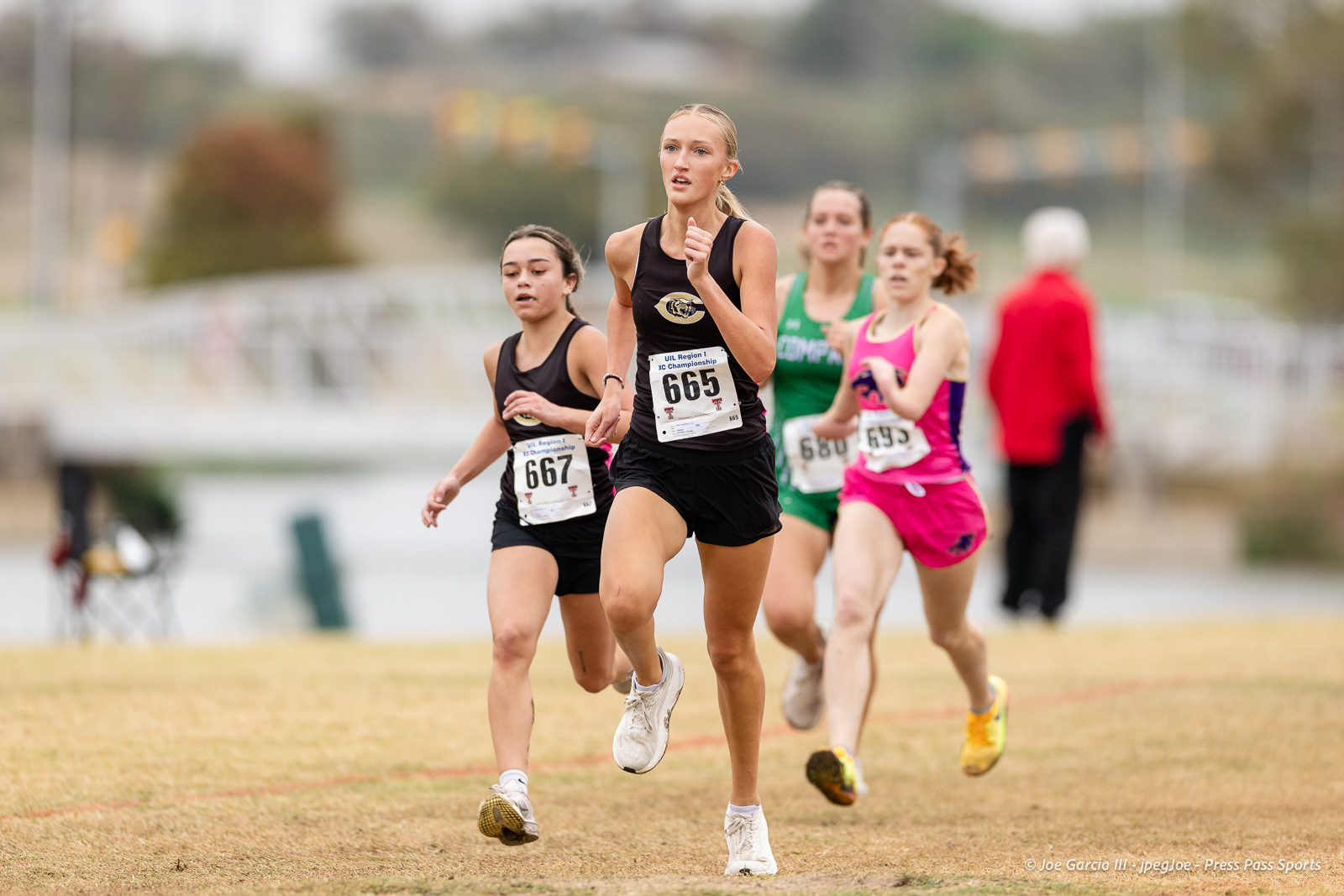 regional-xc-lubbock-2026-jpegJoe-pps-19 | Press Pass Sports Female runners in a cross-country race; lead runner in black with bib 665 sprinting on a grassy field.