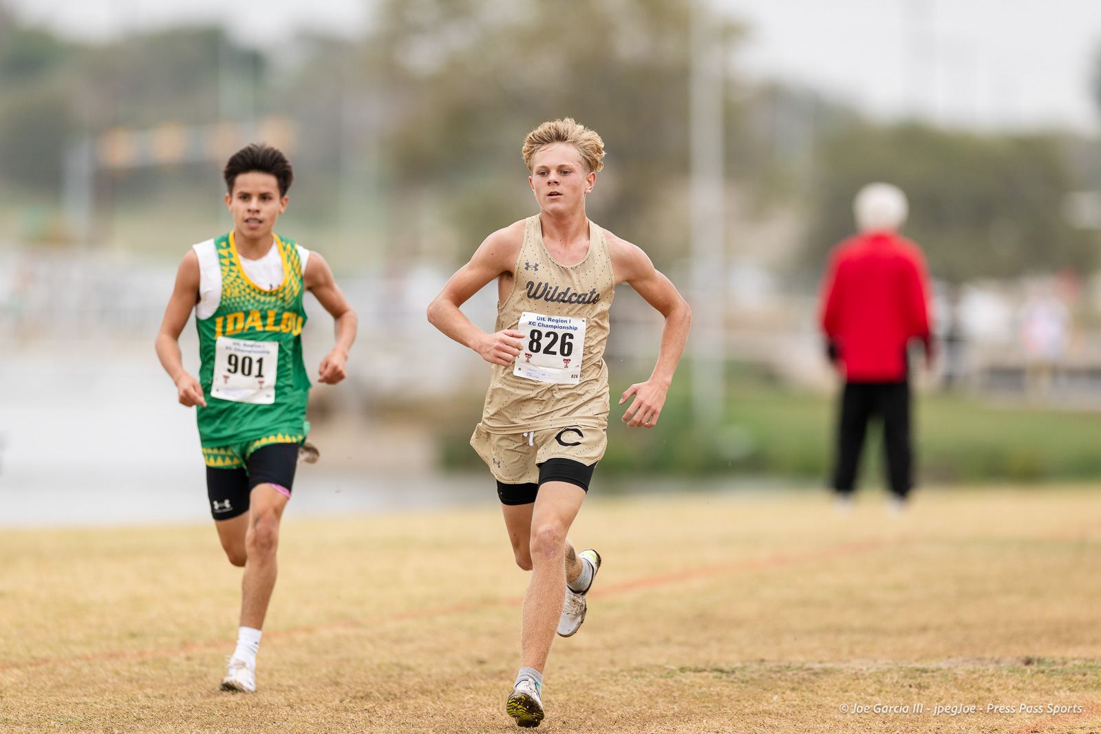 regional-xc-lubbock-2026-jpegJoe-pps-23 | Press Pass Sports Two young runners compete in a cross-country race, front runner in tan Wildcats uniform wearing bib 826; behind him a rival in green with bib 901.
