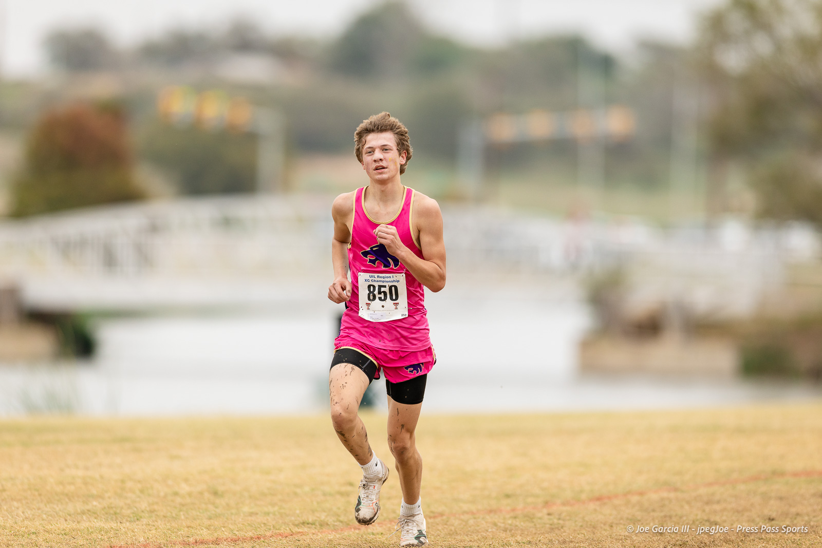 regional-xc-lubbock-2026-jpegJoe-pps-24 | Press Pass Sports Male runner in a pink singlet and shorts, sprinting on a grassy field with a race bib 850.