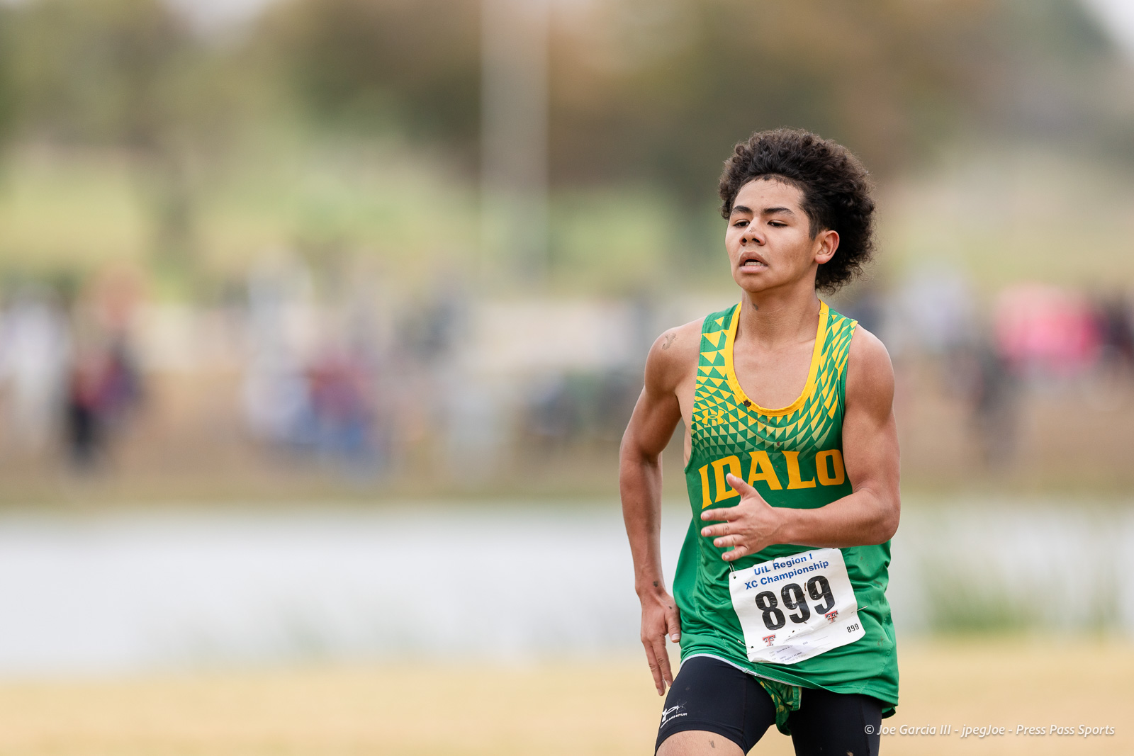 regional-xc-lubbock-2026-jpegJoe-pps-25 | Press Pass Sports Male runner in green and yellow Idaho jersey sprinting on a cross-country course, bib number 899 visible, focused expression.