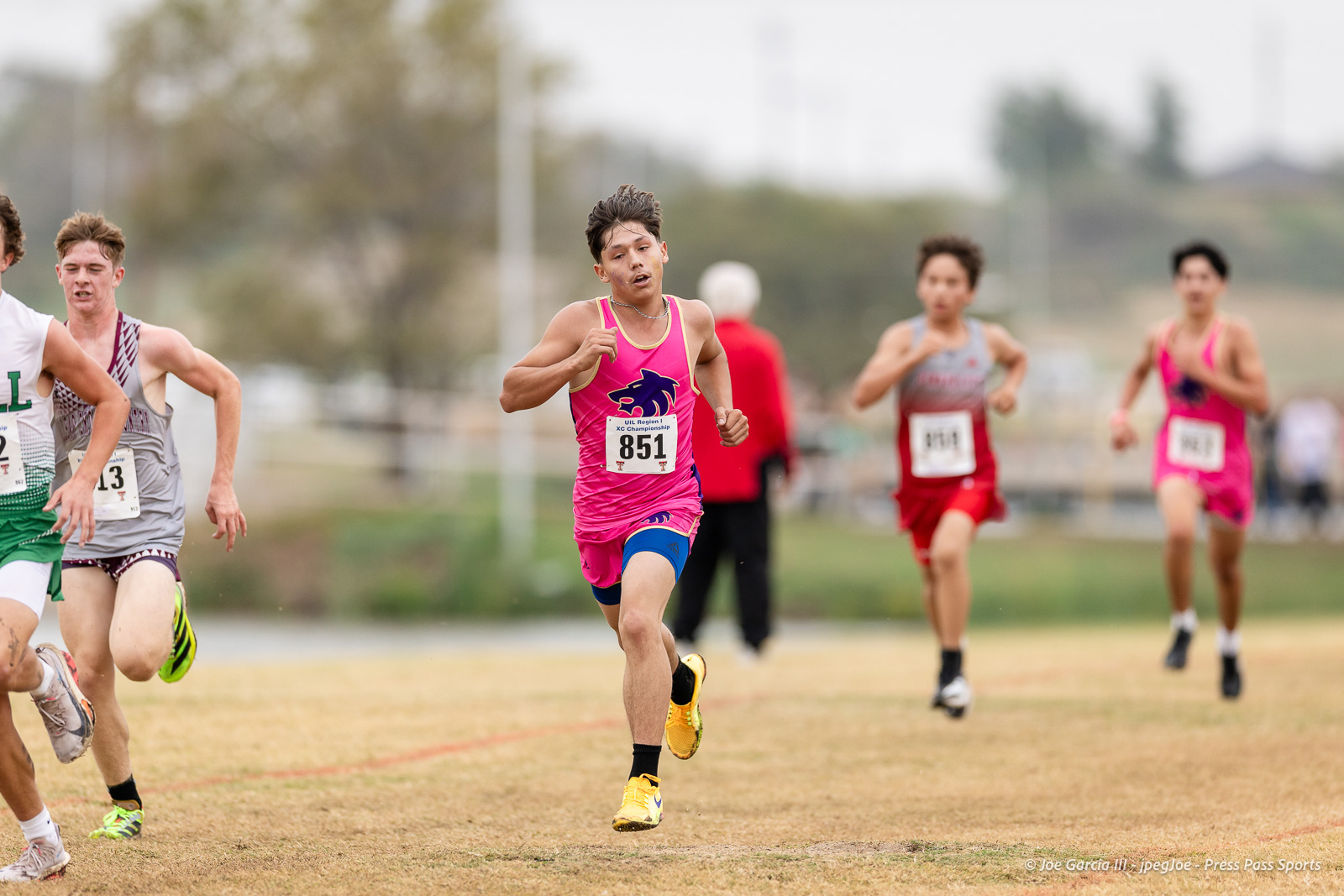 regional-xc-lubbock-2026-jpegJoe-pps-26 | Press Pass Sports Leading male sprinter in a pink uniform (bib 851) on an outdoor track, yellow shoes, others close behind.