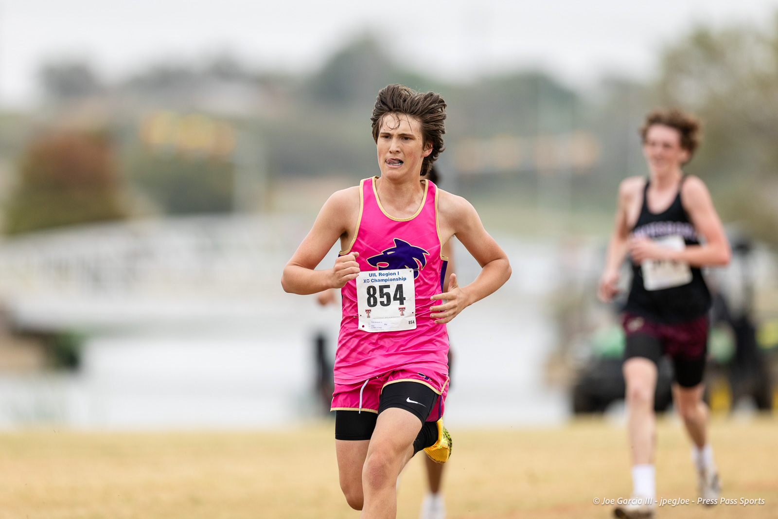 regional-xc-lubbock-2026-jpegJoe-pps-28 | Press Pass Sports Young male runner in a bright pink kit with bib 854 leading a race on a grassy track with a blurred background runner behind him.