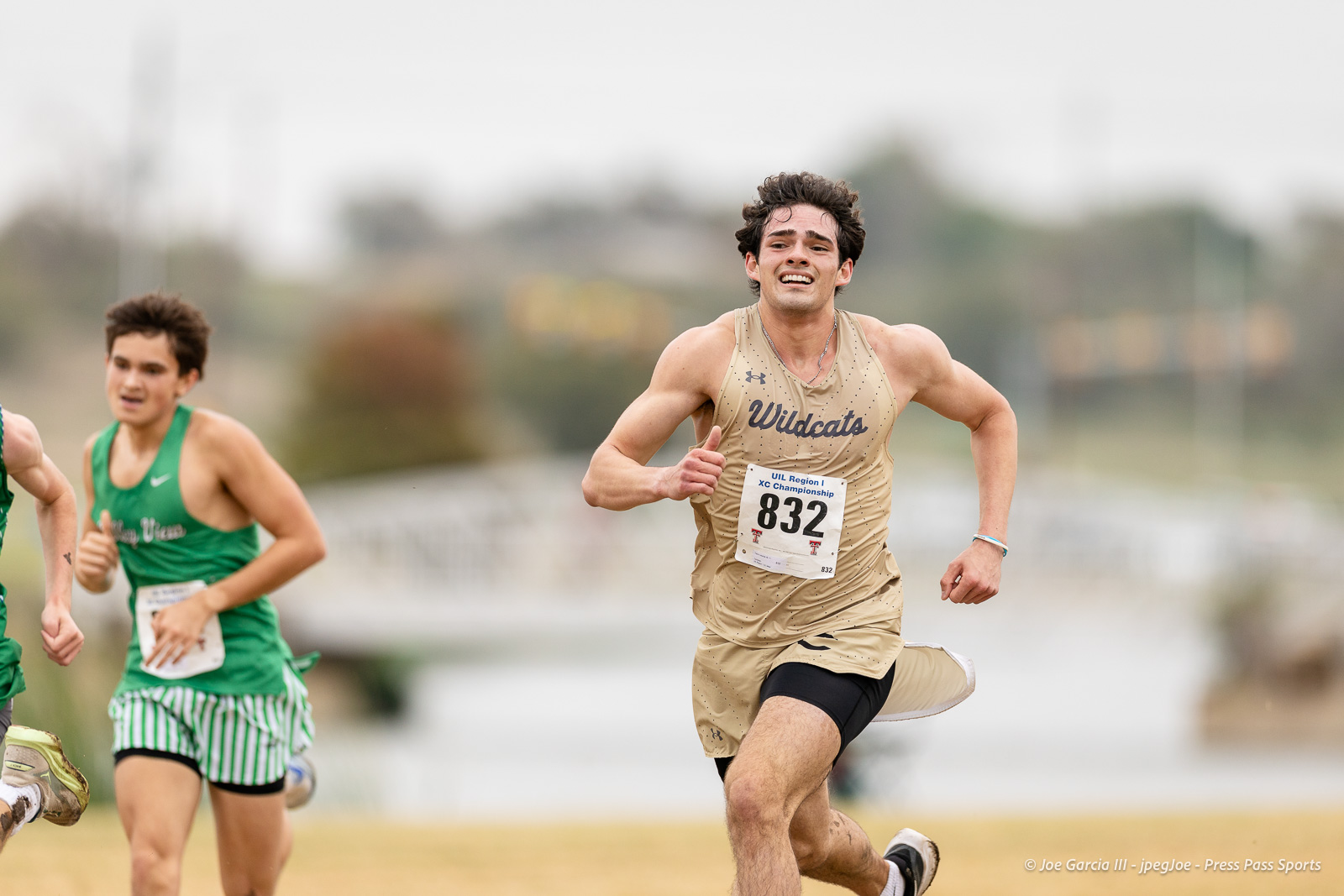 regional-xc-lubbock-2026-jpegJoe-pps-29 | Press Pass Sports Male runner in tan Wildcats uniform with bib 832 leading a race on a track with other competitors nearby in green outfits.