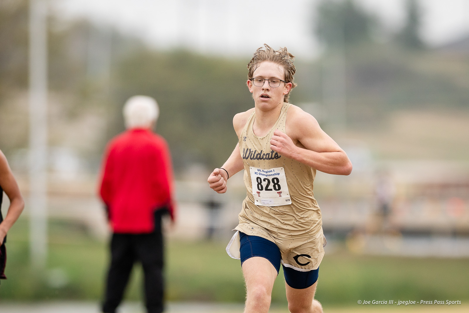 regional-xc-lubbock-2026-jpegJoe-pps-30 | Press Pass Sports Young male cross-country runner in a beige Wildcats jersey with bib 828 sprinting during a race on a blurred field background.