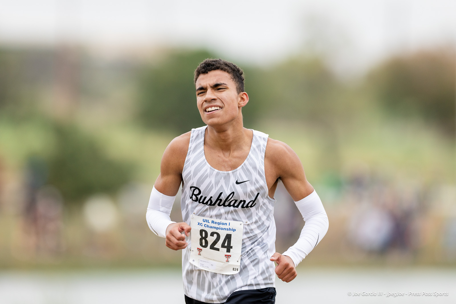regional-xc-lubbock-2026-jpegJoe-pps-31 | Press Pass Sports Male runner wearing a gray patterned jersey with 'Bushland' on front, racing outdoors with bib 824.