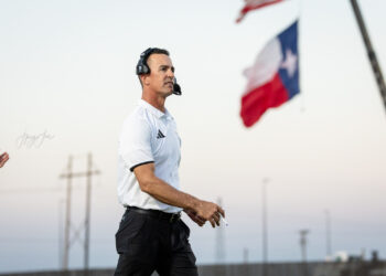 lubbock-christian-vs-bushland-football-jpegJoe-27 | Press Pass Sports Coach wearing a headset and white polo walking on a sports field with a Texas flag in the background on a clear day.