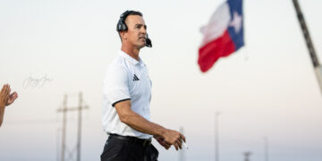 Coach wearing a headset and white polo walking on a sports field with a Texas flag in the background on a clear day.