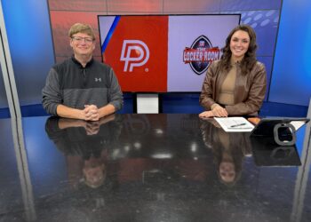 Two TV hosts sit at a glossy desk in a studio, with a large monitor behind them displaying red and light panels and logos.