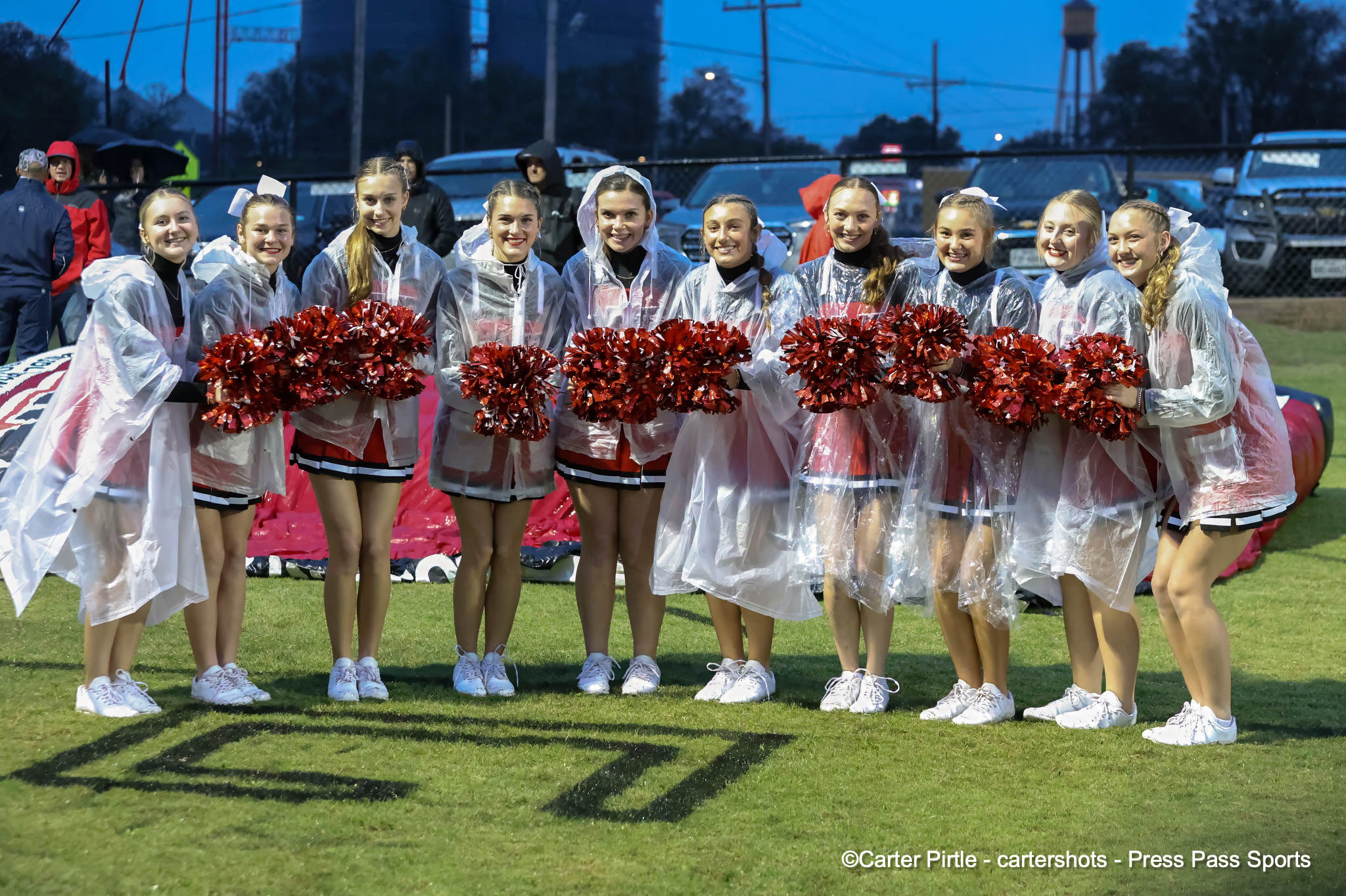 Line of cheerleaders in clear ponchos holding red pom-poms on a grassy field, with a fence and cars in the background.