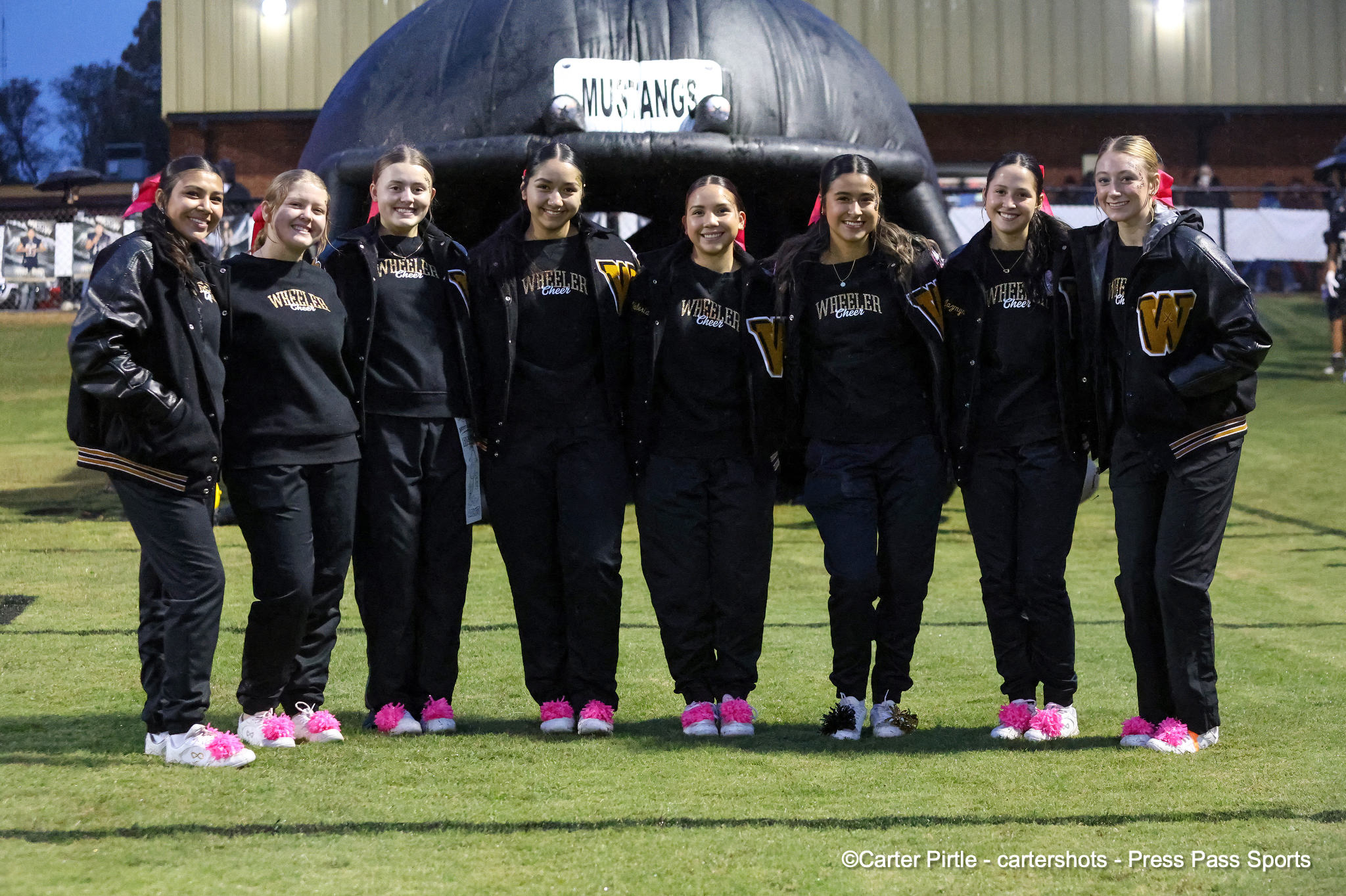 Group of eight cheerleaders in black jackets and pink shoes posing together on a grassy field in front of a large inflatable structure with a 'Mustangs' sign behind them.