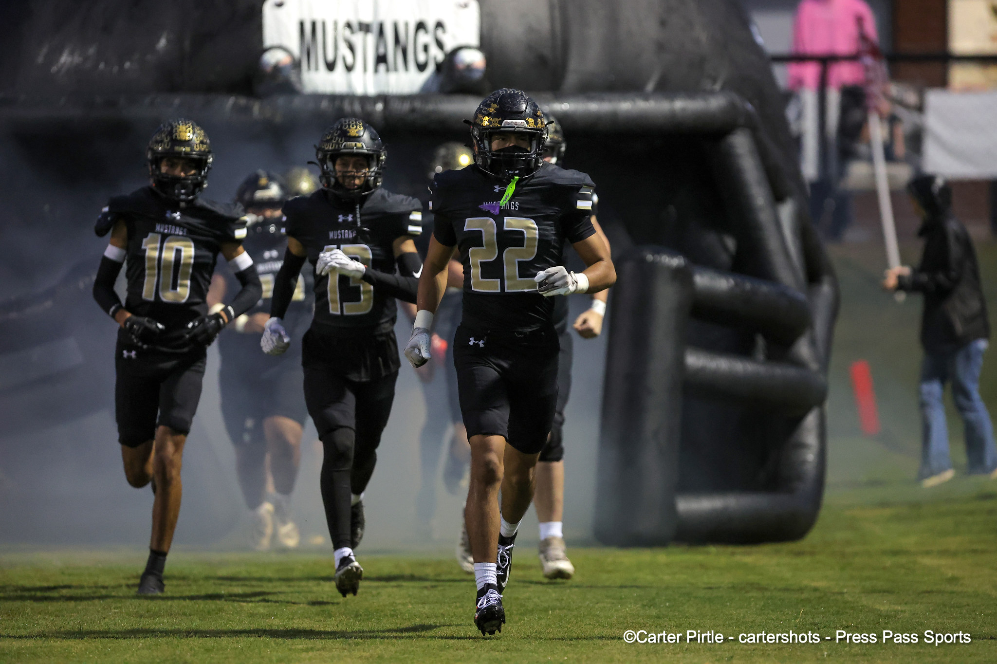 Black-clad football players sprint onto the field through smoke, led by a player wearing number 22 (Mustangs).