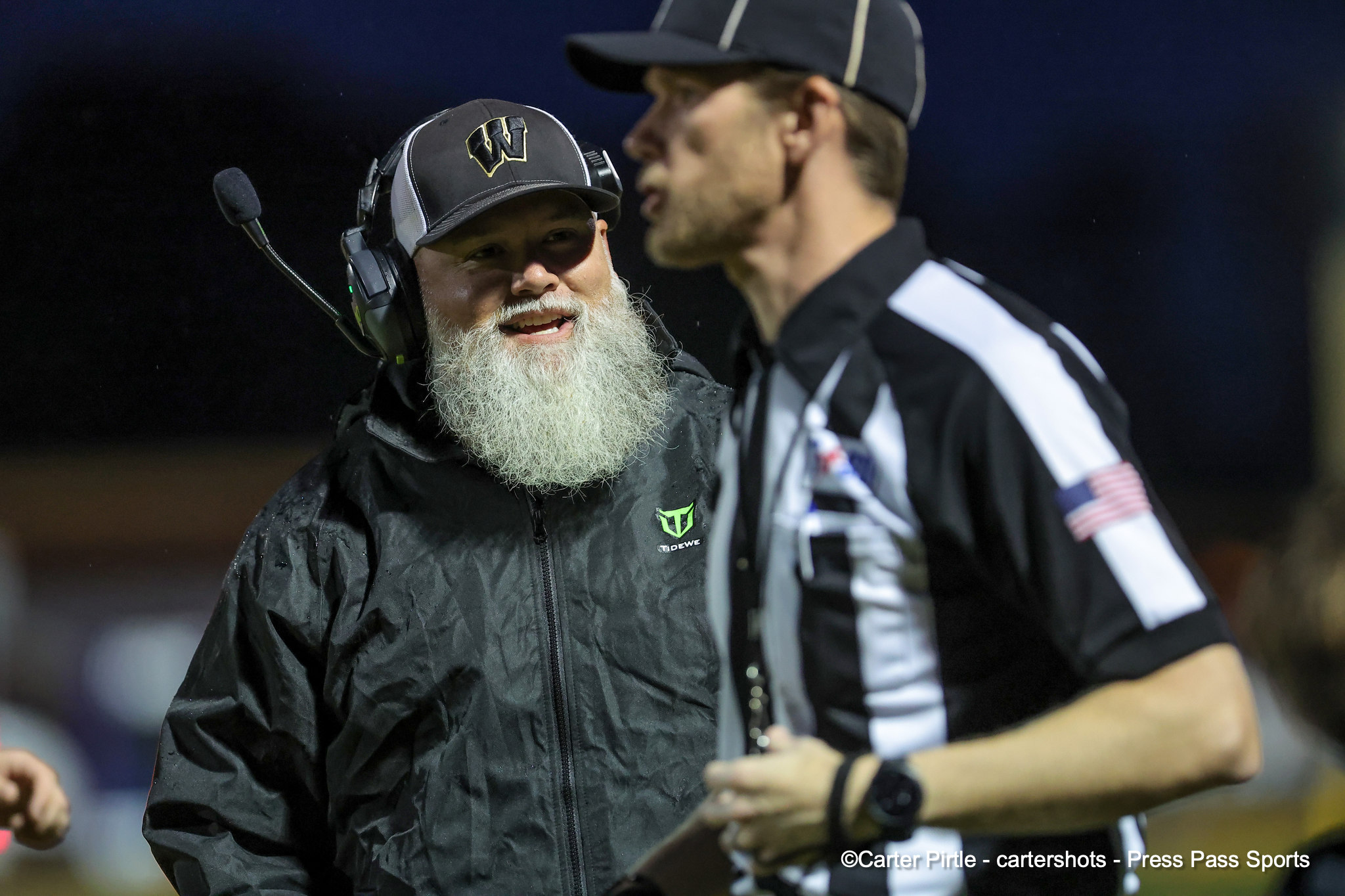 Older coach with a headset and gray beard talks to a referee in a black-and-white striped uniform during a night game.