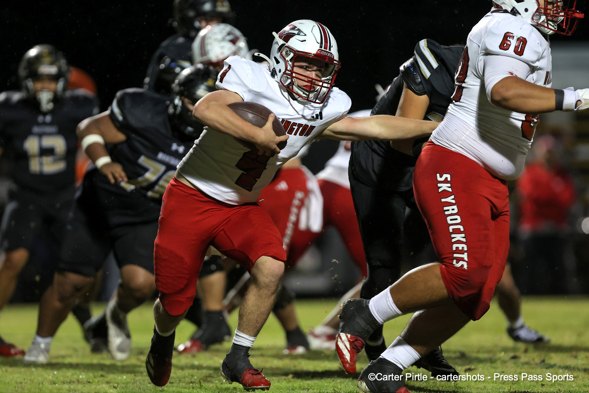 Football player in white and red uniform carries the ball as he runs past defenders during a night game persistently breaking through a tackle.