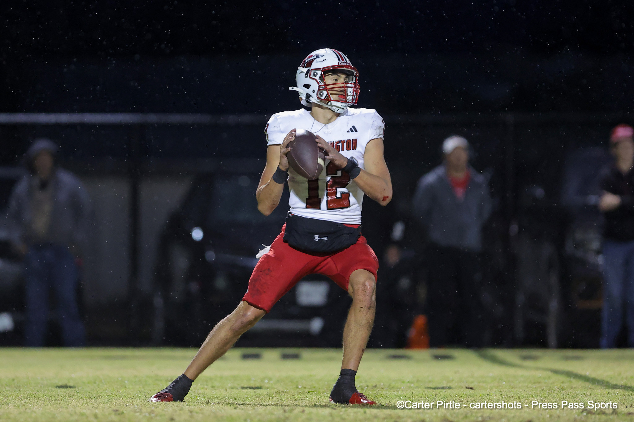 Football player in a white jersey and red shorts runs with a football on a rainy night field, stadium lights in the background.