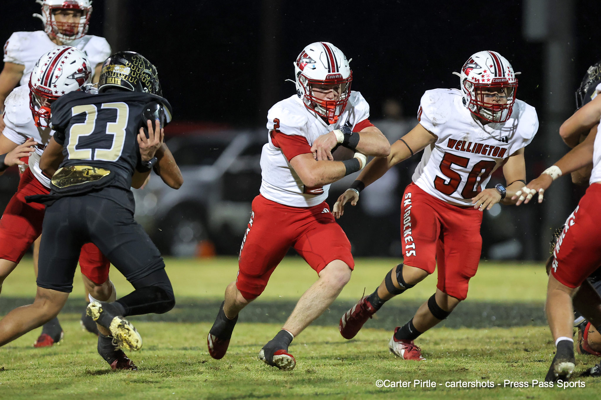 Wellington football players in white and red jerseys sprint on a grass field during a night game, pursuing an opponent in black and gold.