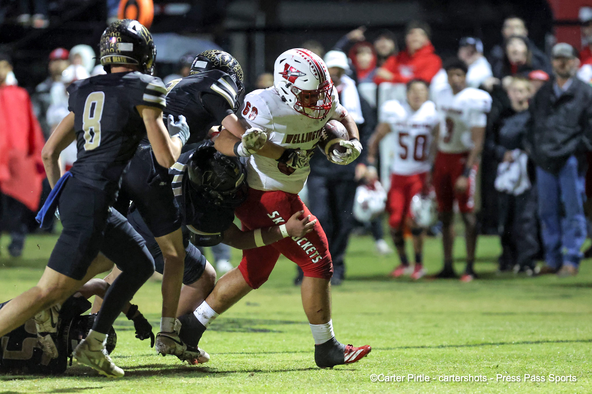A football running back in a white and red uniform carries the ball while defenders in black pursue on a lit football field, with a crowd watching from the stands in the background.
