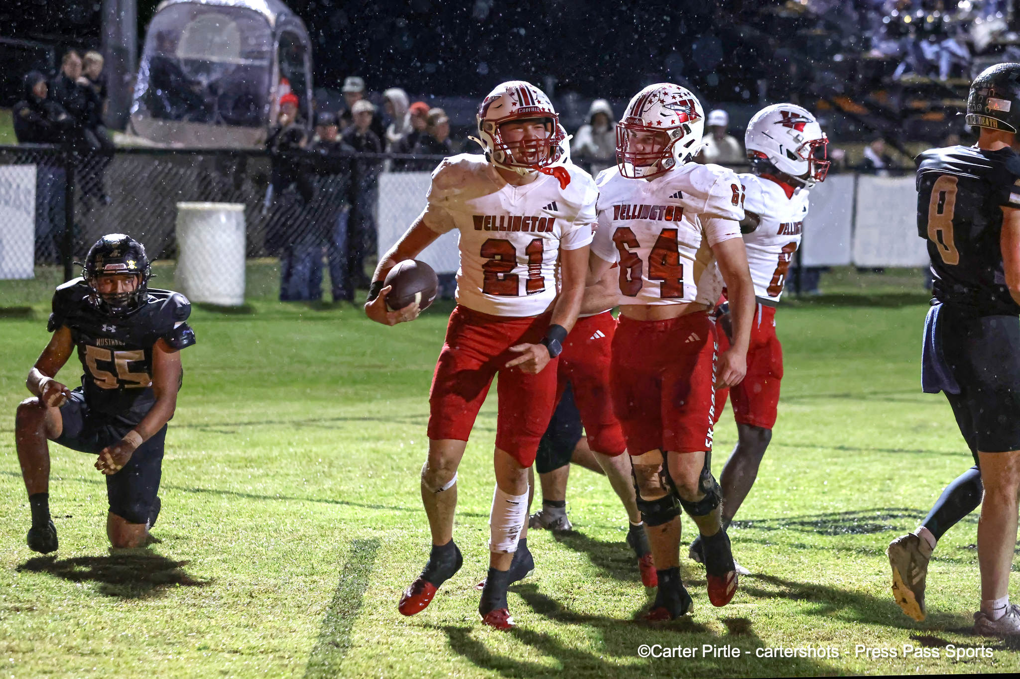 Group of football players in white and red uniforms celebrate on a rain-soaked field at night, a ball visible in one player's hand.