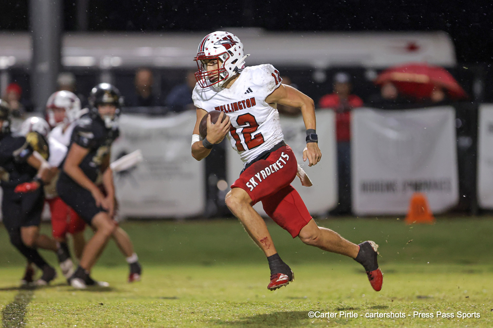 Football player in a white Wellington jersey (#12) runs with the ball during a night game in the rain, his leg muscles tensed as he moves forward.