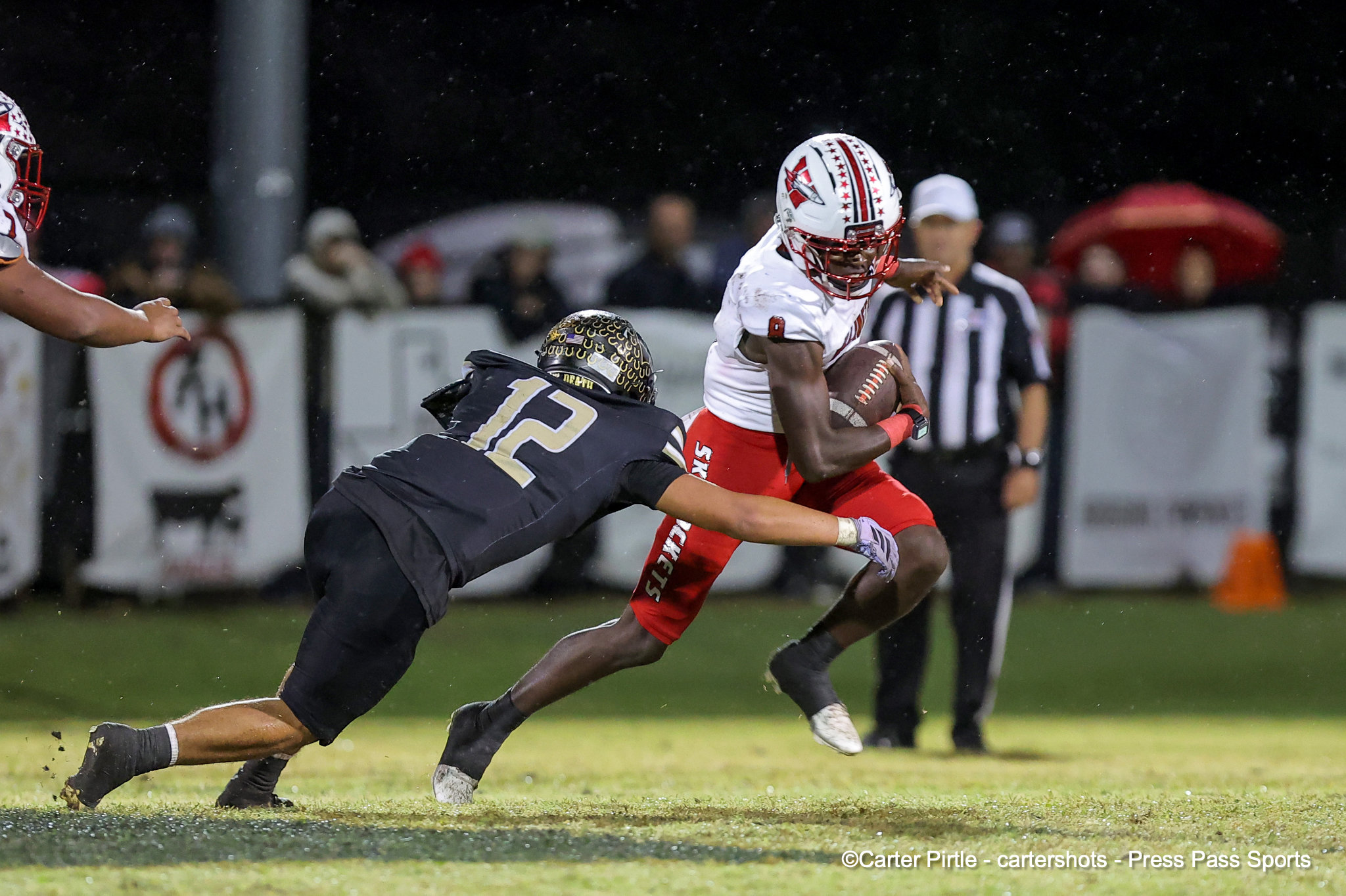 Football player in a white and red uniform runs with the ball while a defender in black tries to tackle him on a rainy night game.