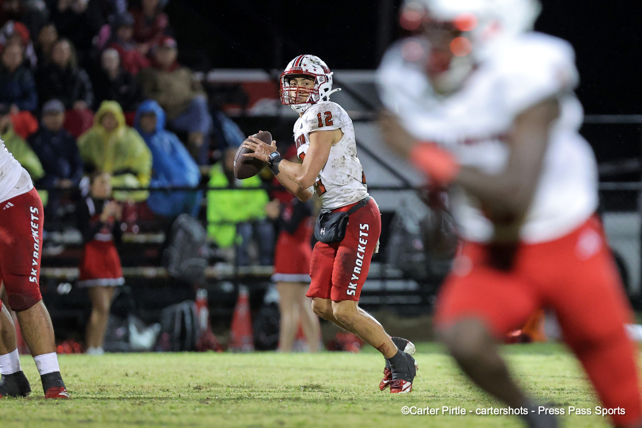 Quarterback in red-and-white uniform running with the football during a night game, crowd in the stands behind him