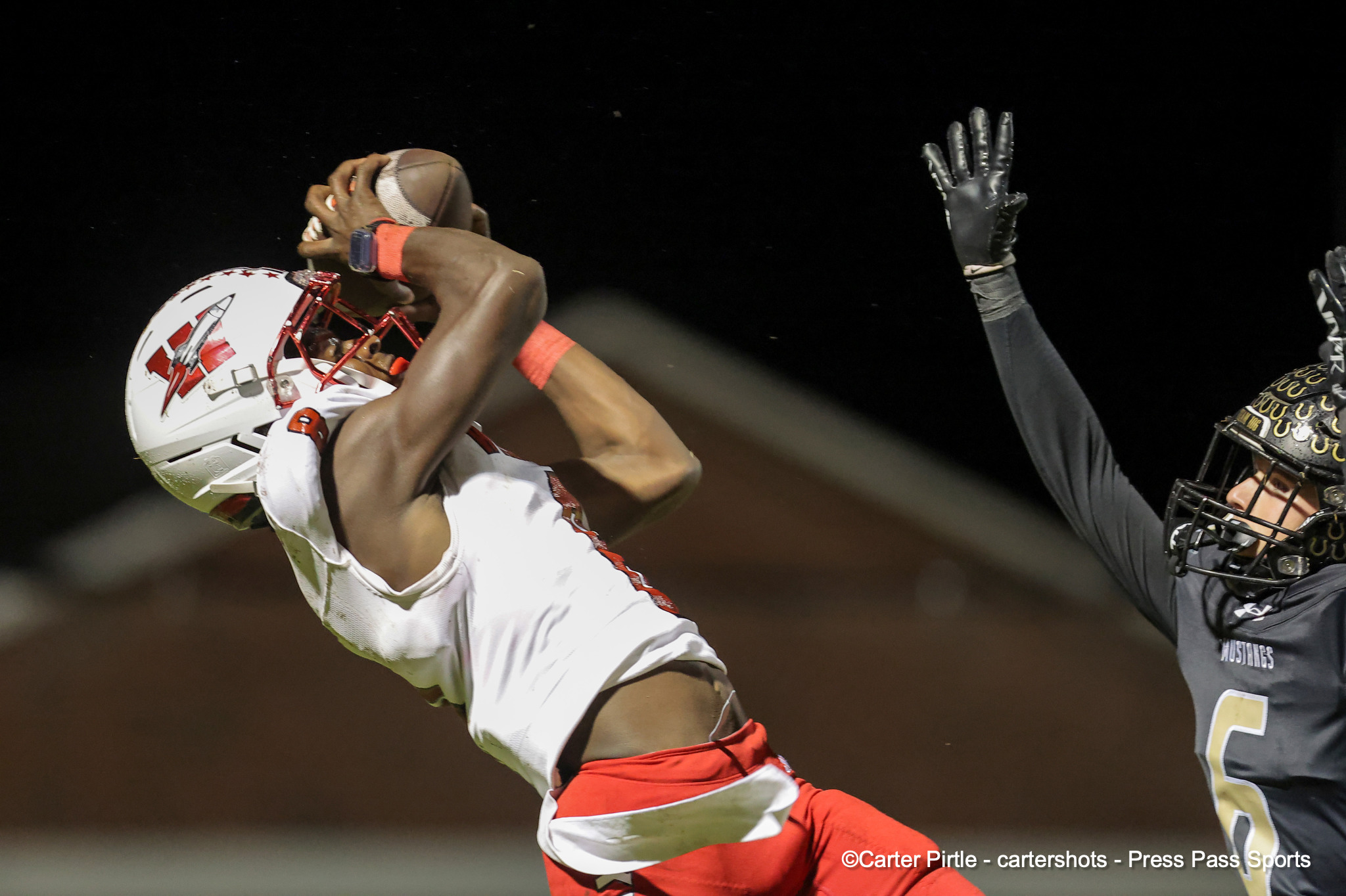 Football player in white and red reaches up to catch a ball as a defender in dark gear extends a gloved hand during a night game.