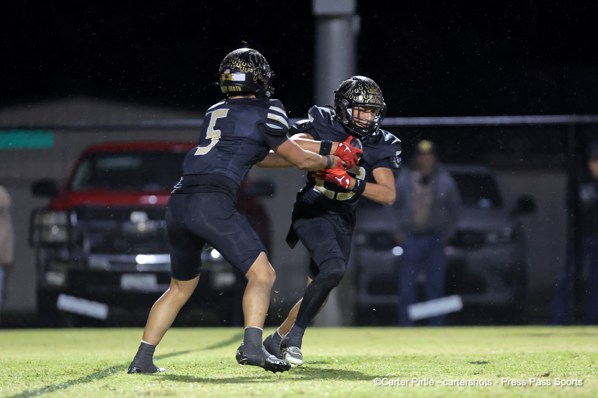 Two football players in dark uniforms collide while tackling on a lit night game in progress on a grassy field.