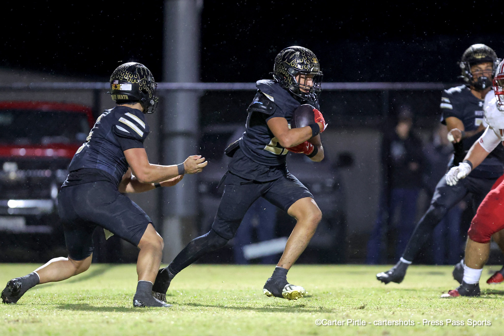 Nighttime football game: a player in a dark uniform runs with the ball as teammates chase in the rain on a grassy field