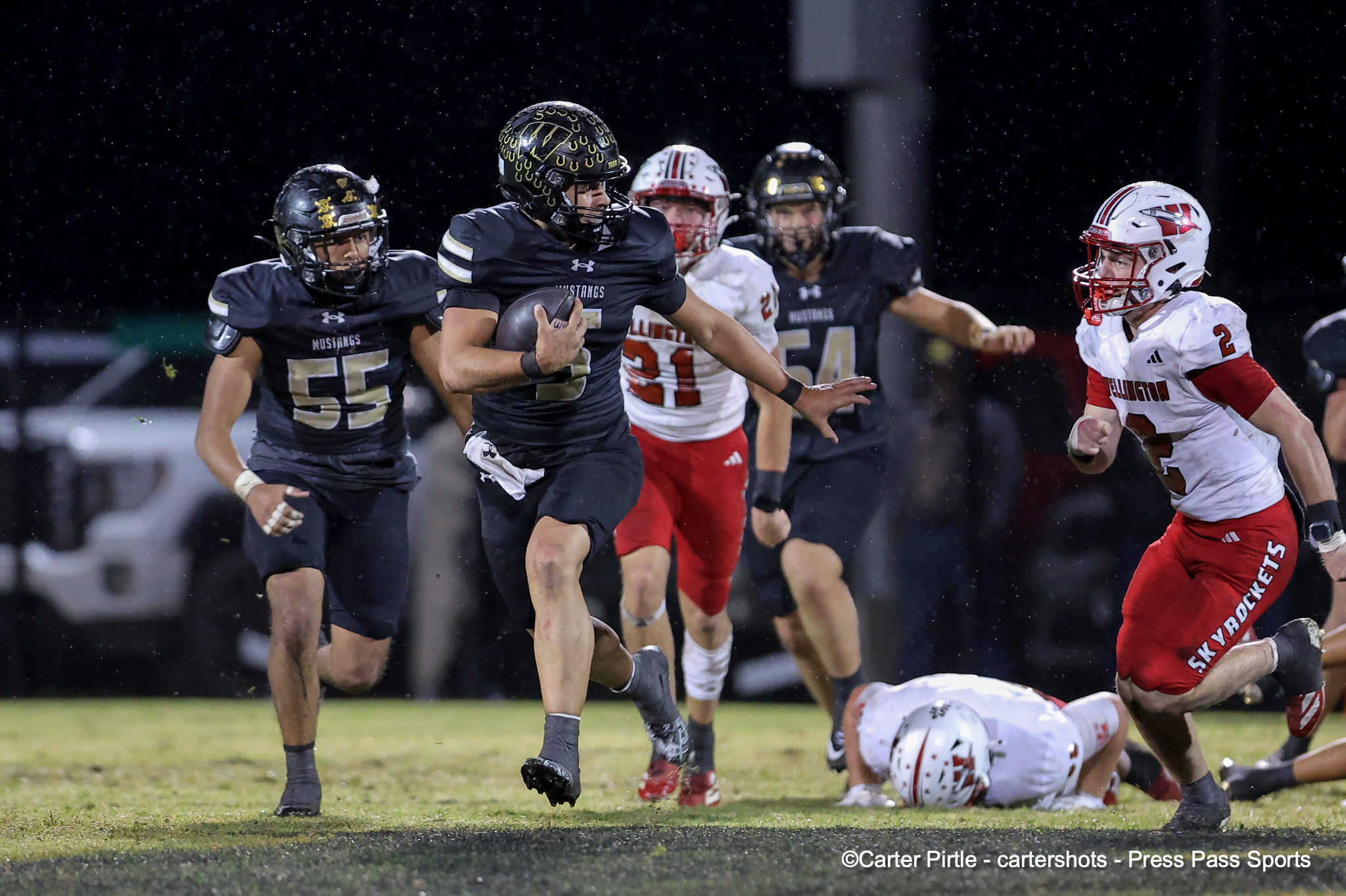 Dark-uniform Mustangs player with ball runs forward while pursued by white-uniform opponents on a snowy night game.