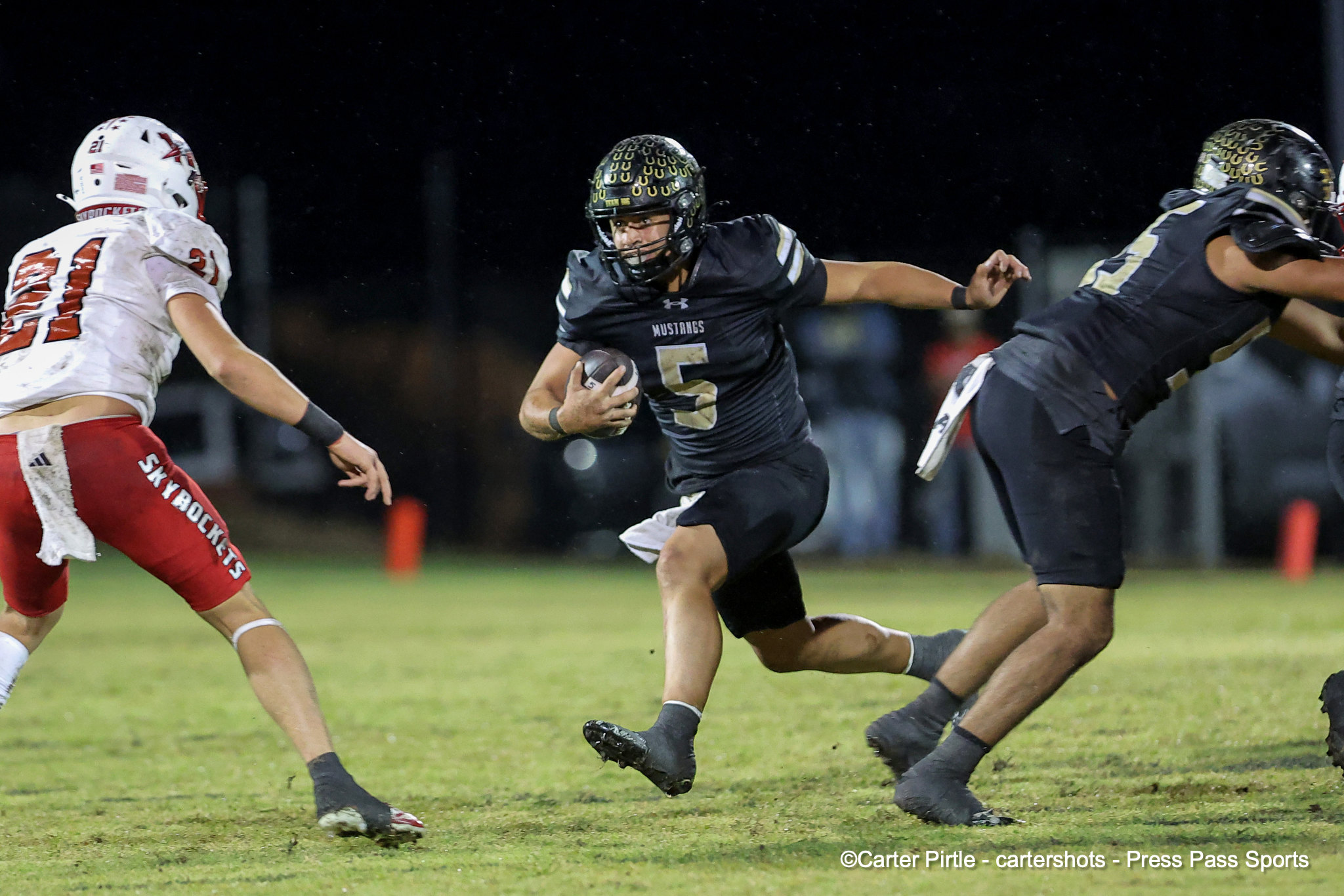 Mustangs running back #5 carries the ball as defenders close in during a nighttime football game on a grassy field.