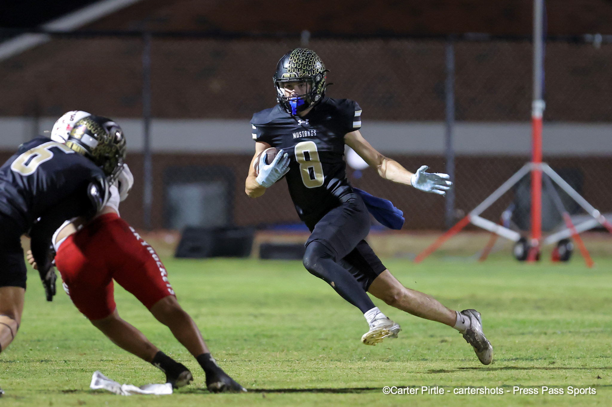 Running back in a black Mustangs uniform (#8) carries the football while sprinting across a grassy field at night as a defender closes in behind him.
