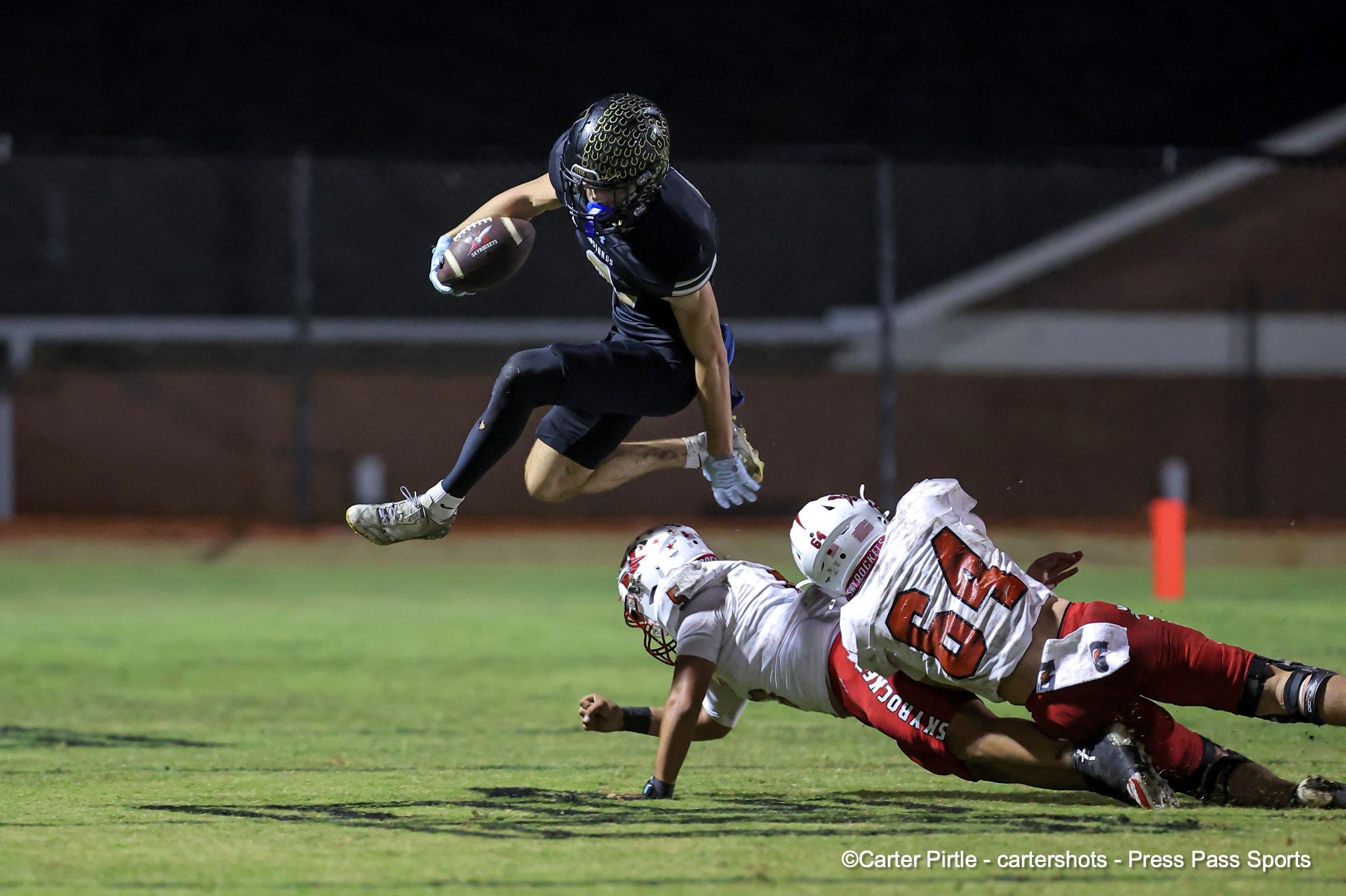 Night football action: a black-uniformed player jumps over a on-the-ground defender to reach the ball in midair under stadium lights.