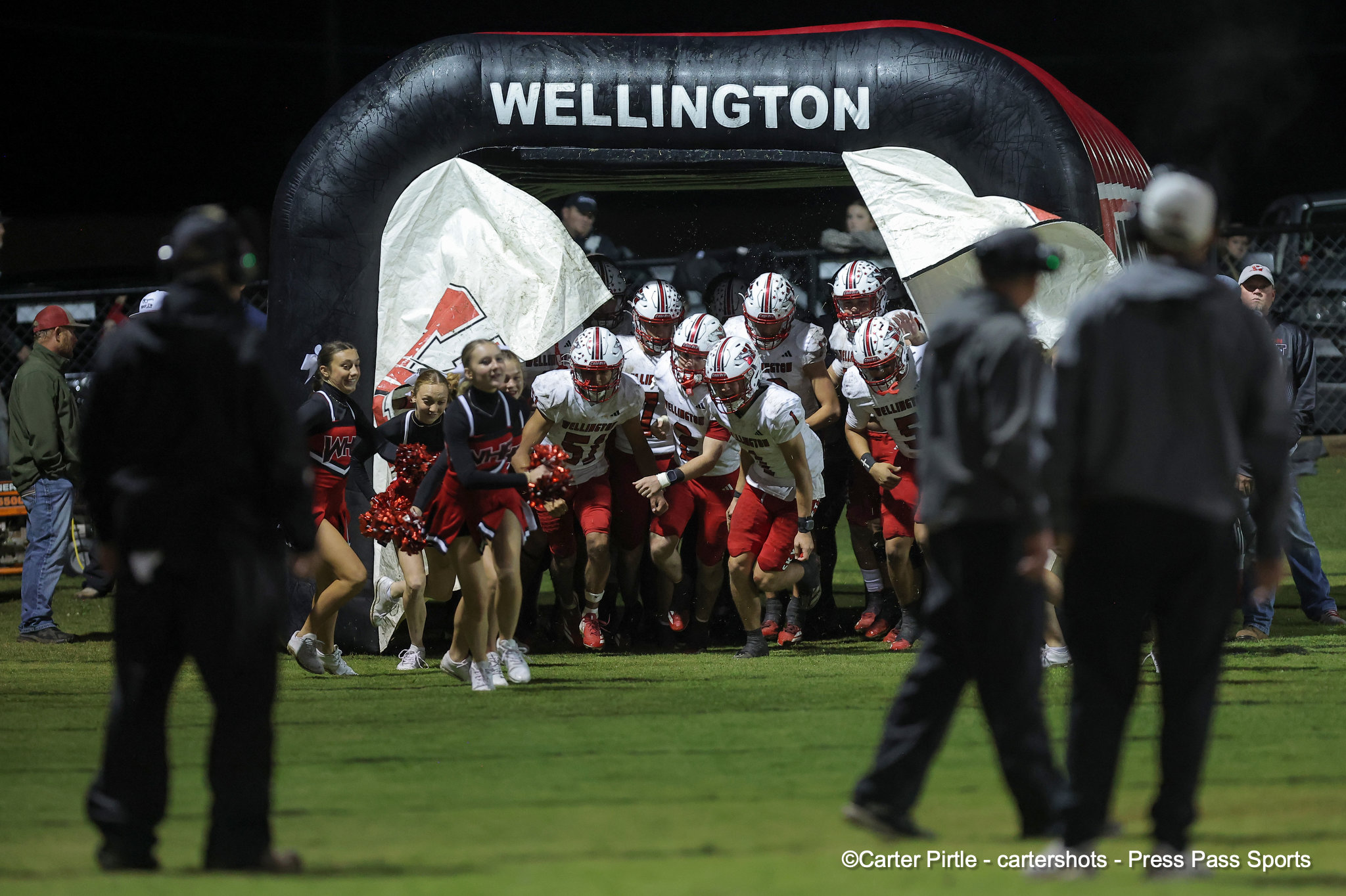 High school football team bursts from a black inflatable tunnel labeled Wellington, sprinting onto the field with cheerleaders nearby at night.